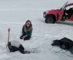 A person kneels in a snow beside a freshly dug hole in the ice.
